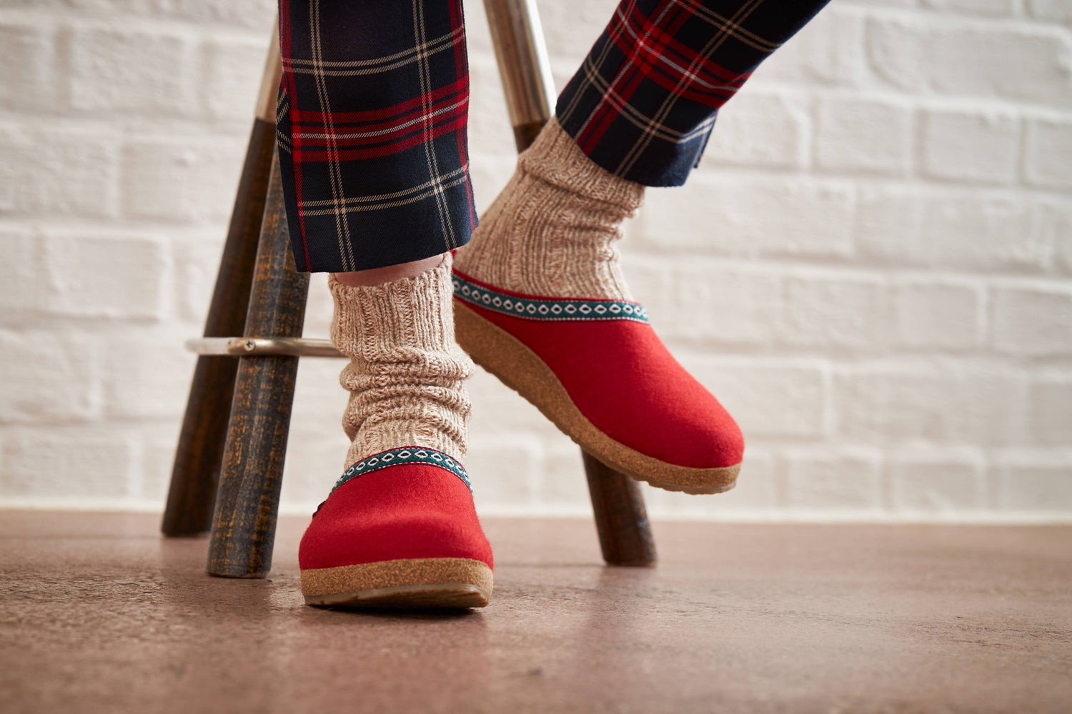 A person wearing red wool clog slipper and thick beige socks sits on a wooden stool, with plaid pants visible, against a white brick wall background.