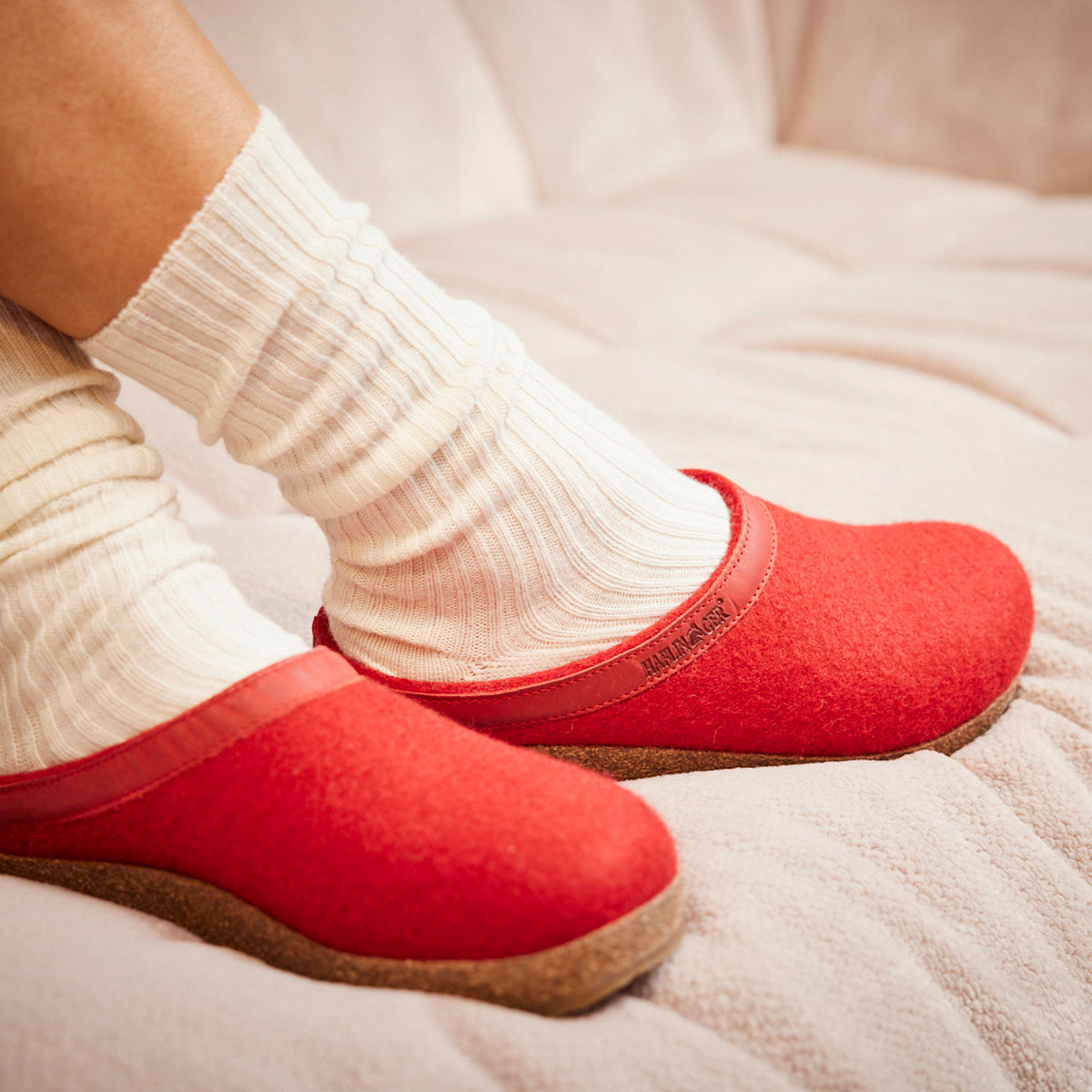 Person wearing white socks and bright red slippers sits on a light-colored cushioned surface.